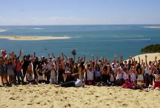 Large group of people with raised arms celebrating on a sandy beach with turquoise sea and sand dunes in the background