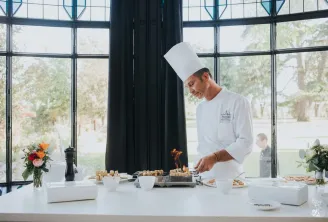 Professional chef in white uniform and tall hat preparing gourmet appetizers at a modern counter with large windows overlooking gardens, with fresh flowers and catering supplies displayed