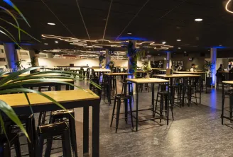 Modern event space at Arkea Arena in Floirac, Bordeaux, featuring high ceilings with decorative lighting, black bar-height tables with wooden tops, black stools, green plants, and people gathered in the background during an evening event.