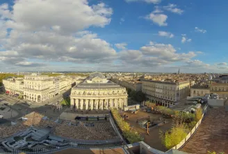 Aerial panoramic view of Bordeaux city center with neoclassical buildings, historic architecture, and urban landscape under a partly cloudy blue sky.