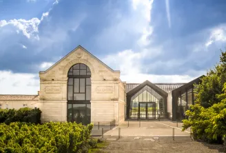 Modern wine cellar building with cream-colored stone facade and large arched window, contemporary glass extension, flanked by green hedges and vineyard buildings under blue sky with clouds