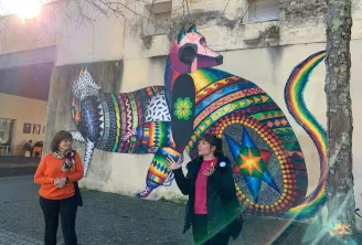 Two women standing in front of a colorful street art mural depicting a stylized animal with geometric patterns and a rainbow tail on a building in Bordeaux.