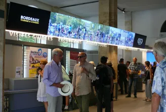 Visitors at the Bordeaux Tourism and Congress Office reception area, with a large digital screen displaying city scenes and information about guided tours and attractions