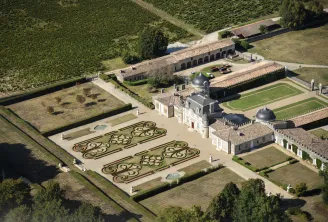 Aerial view of Château de Malle in Bordeaux showing the main residence, formal gardens with ornate parterre patterns, vineyard fields, and stone outbuildings surrounded by manicured hedges and green spaces.
