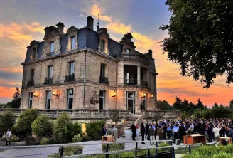 Château Grattequina in Bordeaux at sunset, a grand Second Empire mansion with mansard roof and slate turrets, surrounded by guests at an outdoor evening event, with manicured gardens and tree-lined grounds.