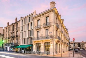 Mercure Bordeaux Gare Atlantic hotel, a beige stone building with classical architecture, ground floor retail spaces with green awning, and ornate wrought iron balconies, photographed at twilight in Bordeaux.