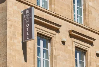 Facade of a historic stone building with a brown hotel sign reading 'HPEC HOTEL' hanging from the wall, featuring regular windows with ornate stone frames and architectural details