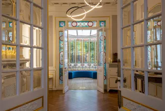 Elegant hallway with ornate double doors opening onto a luxurious room featuring a blue velvet bench, decorative stained glass windows, parquet flooring, and modern circular pendant lights, exemplifying refined Bordeaux interior design.