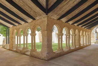 Medieval stone cloister with Romanesque arches and columns beneath wooden ceiling beams, part of the Cordeliers monastery in Bordeaux