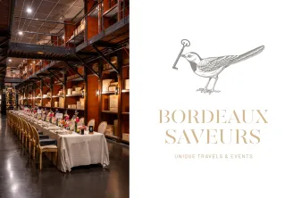 Elegant dining setup at Bordeaux Saveurs with a long white-draped table decorated with pink and red flowers, set in an industrial-style venue with wooden shelving and warm lighting. Bird logo and tagline 'Unique Travels & Events' displayed on the right.
