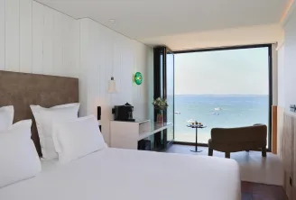 Luxury beachfront hotel bedroom with ocean view, white bed with brown headboard, modern minimalist design, floor-to-ceiling windows opening onto a Mediterranean terrace with boats visible in the water