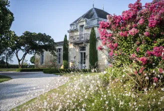 Elegant stone mansion with mansard roof and turret surrounded by blooming pink bougainvillea and white flowers, featuring a gravel courtyard and cypress trees in Bordeaux