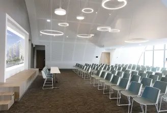 Modern conference room at Hilton Garden Inn Bordeaux Centre with rows of gray chairs, a presentation screen displaying an architectural project, wooden bench seating, and circular pendant lighting fixtures on the ceiling.