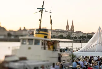 A research vessel with yellow and white hull docked at a Bordeaux waterfront event, with a white tent, crowds of visitors, and the twin spires of a cathedral visible across the river at sunset.
