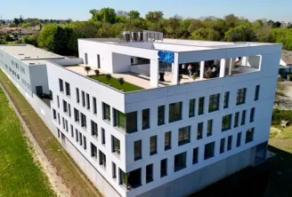 Aerial view of a modern white residential building with multiple floors and a rooftop terrace featuring blue structures, surrounded by green spaces and suburban landscape near Bordeaux.