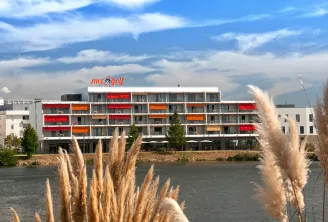 Modern apartment-hotel building with red and orange balconies situated along a waterway, featuring the Mer & Golf signage on the roof, surrounded by contemporary architecture and natural vegetation.