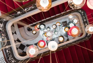 Overhead view of an ornate chandelier with multiple colored glass candle holders in amber, red, and clear tones, suspended in a stairwell with burgundy walls and decorative black railings at Hôtel Villas Foch in Bordeaux.