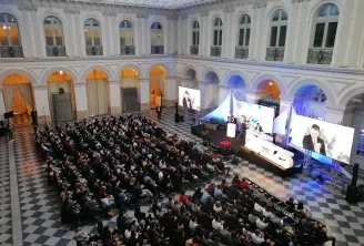 Large conference or gala event in the neoclassical Palais de la Bourse in Bordeaux, with hundreds of attendees seated facing a stage with multiple screens displaying speakers and presentations, ornate architecture with arched windows and columns visible i