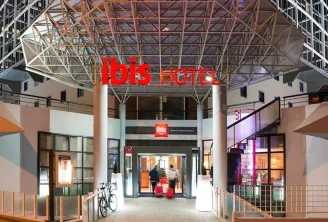 Night view of the Ibis Hotel Bordeaux Centre Mériadeck entrance, featuring a modern architectural canopy with metal framework, illuminated red signage, and contemporary urban setting with surrounding buildings.