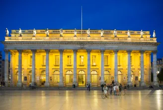 The Grand Théâtre of Bordeaux illuminated at night, featuring a classical facade with Corinthian columns, statues on the roofline, and a flagpole, with people and a cyclist in the plaza below against a deep blue evening sky.