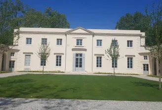 A neoclassical cream-colored manor house with symmetrical facade, multiple windows, central entrance, and triangular pediment, surrounded by manicured lawns and young trees under clear blue sky.