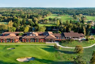 Aerial view of a luxury red-brick golf resort with modern architecture, featuring manicured greens, bunkers, and a clubhouse overlooking a river and forested landscape.