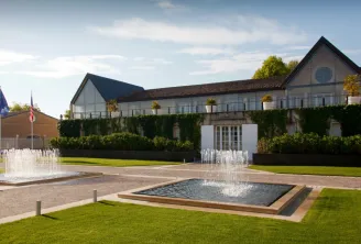 Modern architectural complex with white-walled buildings featuring dark roofs, surrounded by manicured lawns, decorative fountains, and hedges. European flags visible on the left side.