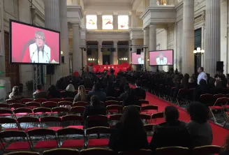 Large conference hall in Bordeaux with classical architecture, red carpet and seating, displaying a speaker on pink-background screens, with attendees seated throughout the ornate space featuring tall columns and natural light from upper windows.