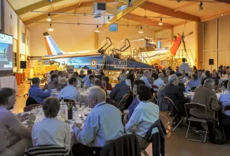 A formal dinner event held inside a hangar at Aérocampus Aquitaine in Bordeaux, with dozens of guests seated at tables in front of a restored military aircraft on display. A presentation screen is visible on the left side of the venue.
