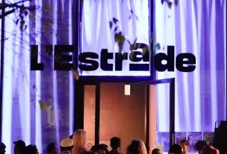 Nighttime exterior view of L'Estrade venue in Bordeaux with illuminated purple and white striped facade, crowds of people gathered at the entrance