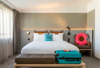 Modern hotel bedroom at Moxy Bordeaux featuring a queen bed with gray upholstered headboard, turquoise pillow with motivational text, white bedding, ambient warm lighting, colorful travel accessories including a pink inflatable donut and turquoise suitcas