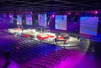 Modern indoor arena with purple and blue lighting, red and white modular stage setup in the center, rows of black seating, large video screens, and professional rigging equipment in Bordeaux.