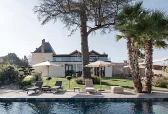 Modern luxury estate with pool, featuring a renovated château tower, contemporary white architecture, palm trees, loungers, and manicured gardens under clear sky