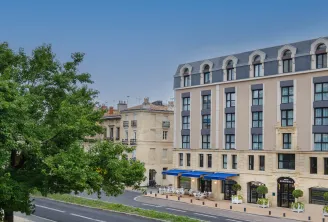 Exterior view of Hôtel Burdigala by Inwood Hotels in Bordeaux, a beige and blue Haussmann-style building with mansard roof, ground-floor café with blue awning, and tree-lined street.
