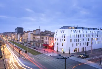 Modern white corner building of Seeko'o Hotel in Bordeaux at dusk, with historic architecture alongside, illuminated streets with light trails from traffic, and tram tracks visible