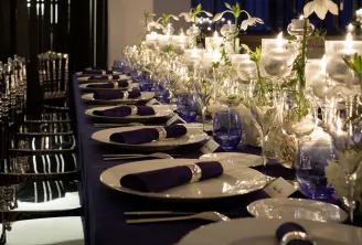 Elegant formal dinner table setting with white plates, dark navy napkins, blue glassware, clear glass vases with white flowers, and candles along a navy tablecloth in a modern event space.