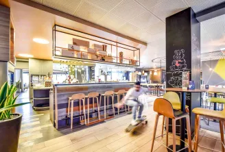 Modern hotel bar and lounge area at Novotel Bordeaux Lac with wooden bar stools, contemporary furniture, open kitchen, and a blurred figure in motion