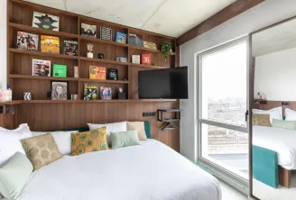 Modern bedroom with wooden wall-mounted shelving unit displaying magazines and decorative items, white bedding with colorful pillows, flat-screen TV, and glass doors opening to a balcony overlooking a city view.
