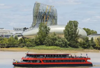 A red and black river cruise boat traveling on the Garonne River in Bordeaux, with the modern La Cité du Vin wine museum building featuring a distinctive tilted glass and metal structure in the background.