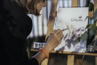 Woman in black jacket writing on a wine tasting note card at an outdoor tasting event in Bordeaux, with vineyard views in the background.