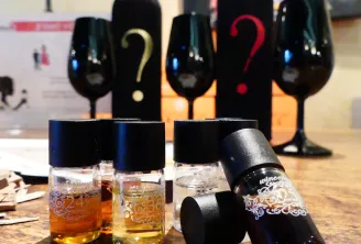 Small bottles of wine with black caps and decorative labels arranged on a wooden surface, with wine bottles displaying question marks in the background, suggesting a wine tasting or selection event in Bordeaux.