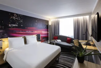 Modern hotel room at Novotel Bordeaux Centre featuring a queen bed with white linens, a gray sofa with red cushions, floor-to-ceiling windows with curtains, and a vibrant cityscape headboard with neon pink and red lights.