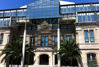 Modern glass and steel structure built atop a historic stone building facade with classical architectural details, flanked by palm trees and white columns at the entrance