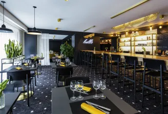 Modern restaurant and bar interior with black tables set with yellow napkins and glassware, a long wooden bar with black stools, illuminated shelving, neon 'hello' sign, pendant lighting, and decorative plants in the Mercure Bordeaux Centre hotel.