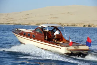 A wooden motorboat named Diletta with red and cream hull moored in shallow waters near a sandy beach, with a large sand dune in the background.