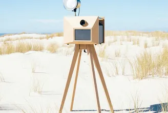 A wooden vintage-style camera on a tripod mounted on a sandy beach with dune grass and ocean in the background