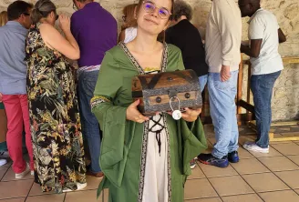 A young woman wearing glasses and a green medieval-style dress holds a wooden treasure chest during a team building activity in Bordeaux, while other participants interact with a stone wall in the background.