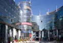 Modern architectural complex featuring a cylindrical glass tower with white framework, surrounded by curved blue-tinted buildings. The central plaza displays signage for 'Centre de Congrès' with decorative red planters and landscaping under clear blue sky
