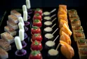 Fine pastry and gourmet appetizers display featuring layered cakes, white cylindrical desserts, red tomato tartlets, black canapés with cream, glazed orange pastries, and herb-topped squares on a black serving board