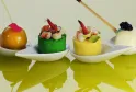 Four gourmet appetizers on white spoon-shaped plates with colored backgrounds: a golden sphere with pink flower, a green mousse with seafood, a yellow mousse with shrimp, and a white panna cotta with berries and paintbrush.